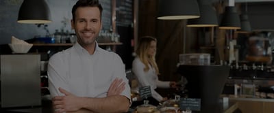 Imprenditore sorridente all'interno del suo locale protetto da sistema di allarme Sector Alarm per attività commerciali, con dispositivi di sicurezza visibili in primo piano