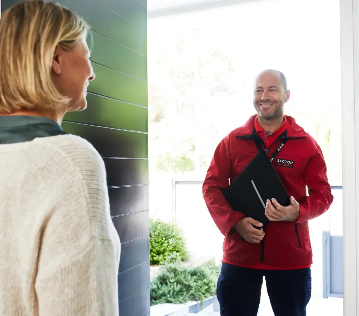 Tecnico Sector Alarm in divisa rossa accoglie con il sorriso una cliente alla porta di casa, pronto per l’installazione del sistema
